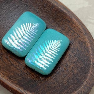 May include: Two teal blue glass tiles with a white fern design. The tiles are iridescent and have a glossy finish.