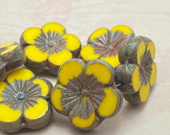 A group of Czech glass hibiscus flower beads in bright yellow with grey picasso finish details displayed on a plain pale.