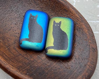 Two laser etched Czech rectangle beads with black cat silhouettes on blue and yellow dichroic backgrounds, shown in a wooden.