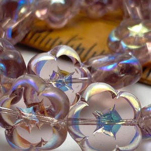 Puede incluir: Primer plano de cuentas iridiscentes en forma de flor. Las cuentas son de color rosa suave con un diseño en forma de estrella en el centro. Las cuentas tienen un efecto brillante, similar al arcoíris. Las cuentas están ensartadas en un alambre fino.
