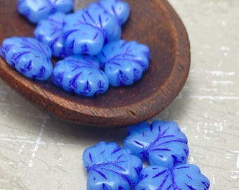 Czech glass maple leaf beads in bright blue with purple wash, displayed in a wooden dish and on a pale textured background.
