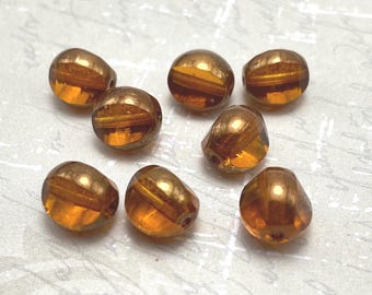Seven topaz orange Czech glass beads with gold detail arranged in a wooden bowl and on a white textured surface.