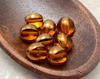 Seven topaz orange Czech glass beads with gold detail arranged in a wooden bowl and on a white textured surface.
