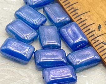 May include: A collection of ten rectangular, iridescent blue beads. Each bead has rounded edges and a glossy finish. The beads are scattered on a light-colored surface, with a wooden ruler in the background.
