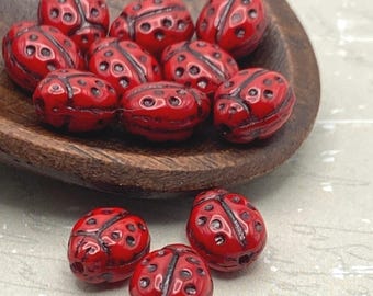 Twelve red Czech glass ladybird beads with black spots displayed in a wooden dish and on a light surface.