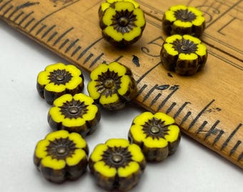 Ten Czech glass hibiscus flower beads in bright yellow with dark brown picasso finish, scattered on a wooden ruler for scale.