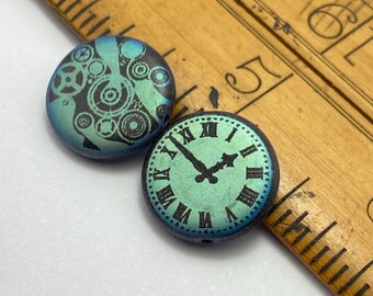 Two matte blue-green laser etched coin beads with steampunk designs on a white background beside a wooden bowl.