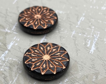 Two black coin beads with a copper-coloured origami flower pattern on a plain textured background.
