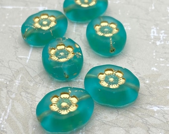 Six matte turquoise green Czech oval flower beads with gold-washed centres, arranged on a pale textured background.