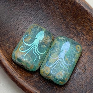 May include: Two rectangular glass beads with a teal blue and gold iridescent finish. Each bead features a white silhouette of an octopus.