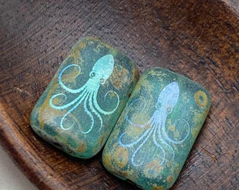 Two turquoise Picasso laser etched Czech glass octopus beads, 18x12mm, on a dark wooden tray.