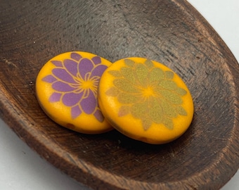 Two matte orange Czech glass coin beads with laser etched flower designs in purple, resting in a dark wooden dish.