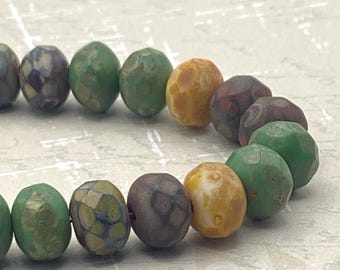 Eighteen Czech glass rondelle beads in green, brown and gold tones with mottled finishes arranged in two rows on a pale.