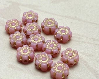 Twelve baby pink Czech glass flower beads with gold wash detail, arranged on a cream coloured linen background.