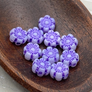 May include: A collection of ten purple glass flower beads. The beads are white with a purple swirl design.