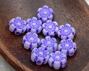 Twelve Czech glass flower beads in white with purple violet wash, arranged in a wooden bowl on a white fabric background.