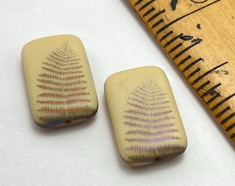 Two ivory cream laser etched Czech rectangle beads with fern leaf pattern shown next to a wooden ruler for scale.