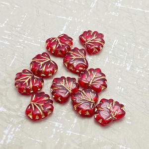 May include: A group of red glass leaf beads with gold accents. The beads are scattered on a white textured surface.