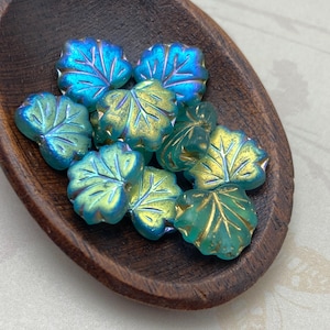 Puede incluir: Un conjunto de cuentas iridiscentes en forma de hoja en una cuchara de madera. Las cuentas tienen una mezcla de tonos azules, verdes y dorados, con vetas detalladas. La cuchara de madera marrón oscuro contrasta con las coloridas cuentas.