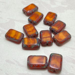 May include: A collection of rectangular beads with rounded edges. The beads have a translucent orange centre with red and teal edges. The beads are arranged on a light-coloured surface.