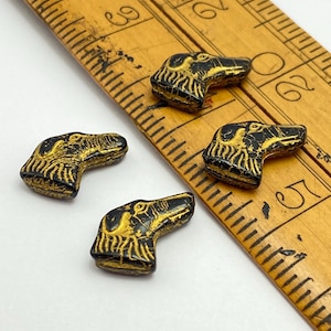 May include: Four black and gold dog head beads. The beads are detailed with gold accents and are positioned near a wooden ruler. The beads are likely for jewelry making or crafting.