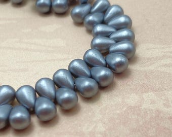 Approximately 50 matte pearl silver grey Czech glass tiny drop beads arranged in curved rows on a plain cream background.