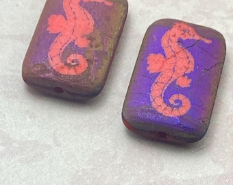 Two Czech rectangle beads with etched seahorse design in purple and orange on a plain background.