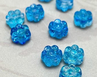 Eleven Czech glass Hawaii flower beads in shades of bright blue and turquoise with AB finish, scattered on a light grey.