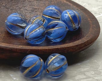 Eight periwinkle blue melon beads with gold wash stripes sit in a dark wooden spoon, with two more beads beside it on white.
