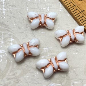 Op de afbeelding: Vier witte glazen vlinder kralen met koperen accenten. De kralen zijn ongeveer 2,5 cm lang.