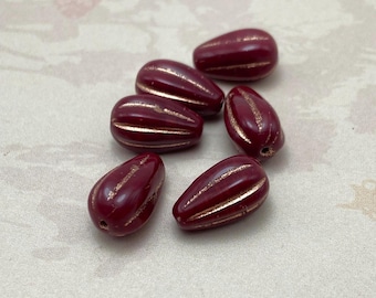 Six burgundy Czech glass melon beads with copper wash detail arranged on a plain beige background.