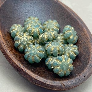 May include: Teal flower-shaped beads with gold detailing, presented in a dark wooden scoop. These textured beads are ideal for jewellery making or craft projects. Each bead has a unique floral design, perfect for adding detail.
