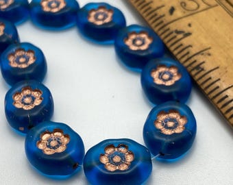 Matte azure blue Czech oval flower beads with copper wash, arranged in a curve next to a ruler for scale.