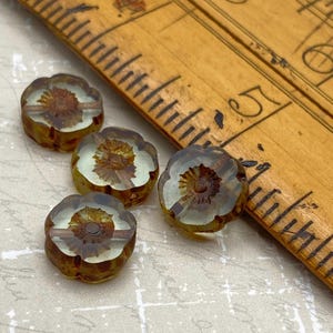 May include: Four flower-shaped beads with a brown centre and petals. The beads are on a light-coloured surface next to a wooden ruler. The beads have a vintage, antique appearance.