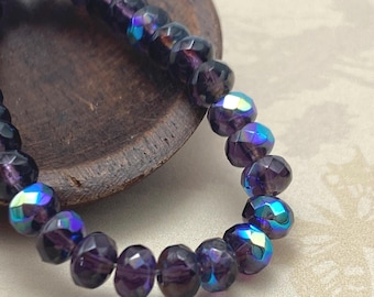 Purple Pansy Czech glass rondelle beads with blue and green AB finish, arranged in a strand on a wooden dish against a cream.