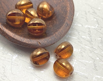 Seven topaz orange Czech glass beads with gold detail arranged in a wooden bowl and on a white textured surface.