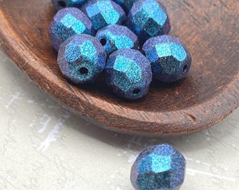 Nine teal blue chameleon Czech glass beads grouped in a wooden bowl with one bead on white cloth below.