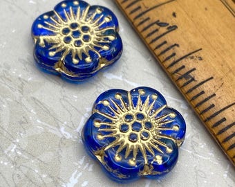 Two sapphire blue Czech glass anemone flower beads with gold details next to a wooden ruler showing scale.