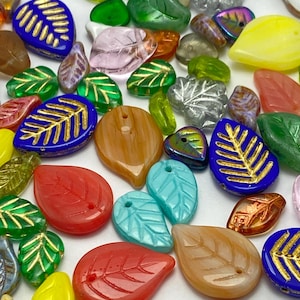 Könnte beinhalten: Eine Sammlung farbenfroher, blattförmiger Perlen in verschiedenen Größen und Farbtönen. Die Perlen bestehen aus Glas und weisen eine Vielzahl von Farben auf, darunter Blau, Grün, Rot, Gelb und Braun. Einige Blätter haben goldene Akzente.