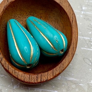 May include: Two turquoise glass beads with gold stripes. The beads are teardrop shaped and have a glossy finish.