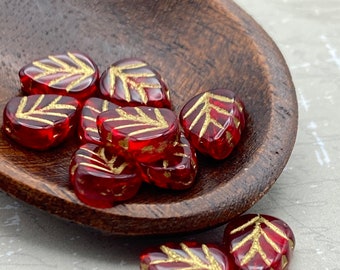Czech glass mint leaf beads in ruby red with gold wash are displayed in a wooden dish and on a pale surface.