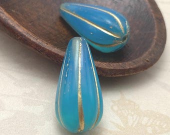 Two matte turquoise Czech glass melon drop beads with gold wash stripes, displayed on a dark wooden dish.