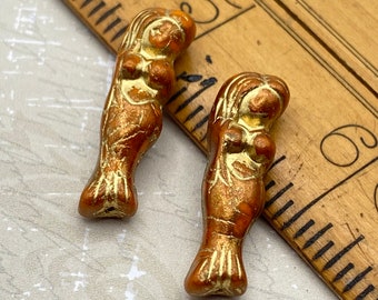 Two burnt orange Czech glass mermaid beads with gold wash finish, shown on a wooden ruler for scale.