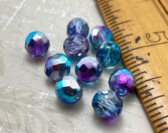 Ten Czech glass faceted beads in shades of blue and purple with iridescent finishes, shown next to a wooden ruler for scale.