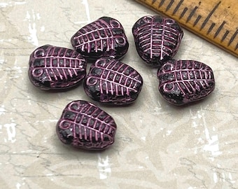 Six black and pink trilobite beads scattered on a plain background next to a wooden ruler for scale.
