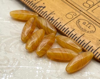Eight golden orange marble Czech glass long oval spindle beads next to a wooden ruler for scale.