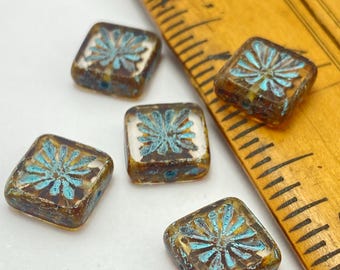 Five Czech glass square beads with turquoise starburst design on bronze background next to ruler for scale.