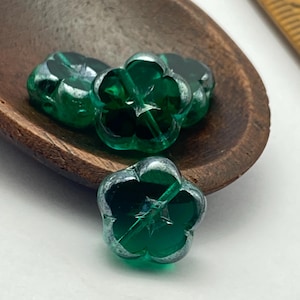 May include: Close-up of four green flower-shaped beads with a silver-colored rim, resting in a wooden scoop. The beads have a translucent quality, and the scoop is a dark brown color.