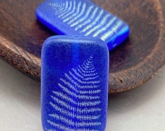 Two matte blue Czech rectangle beads with laser etched fern leaf design displayed on a dark wooden surface.