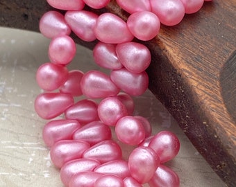 Matte pearl baby pink Czech glass tiny drop beads arranged in a loose strand on a wooden surface.
